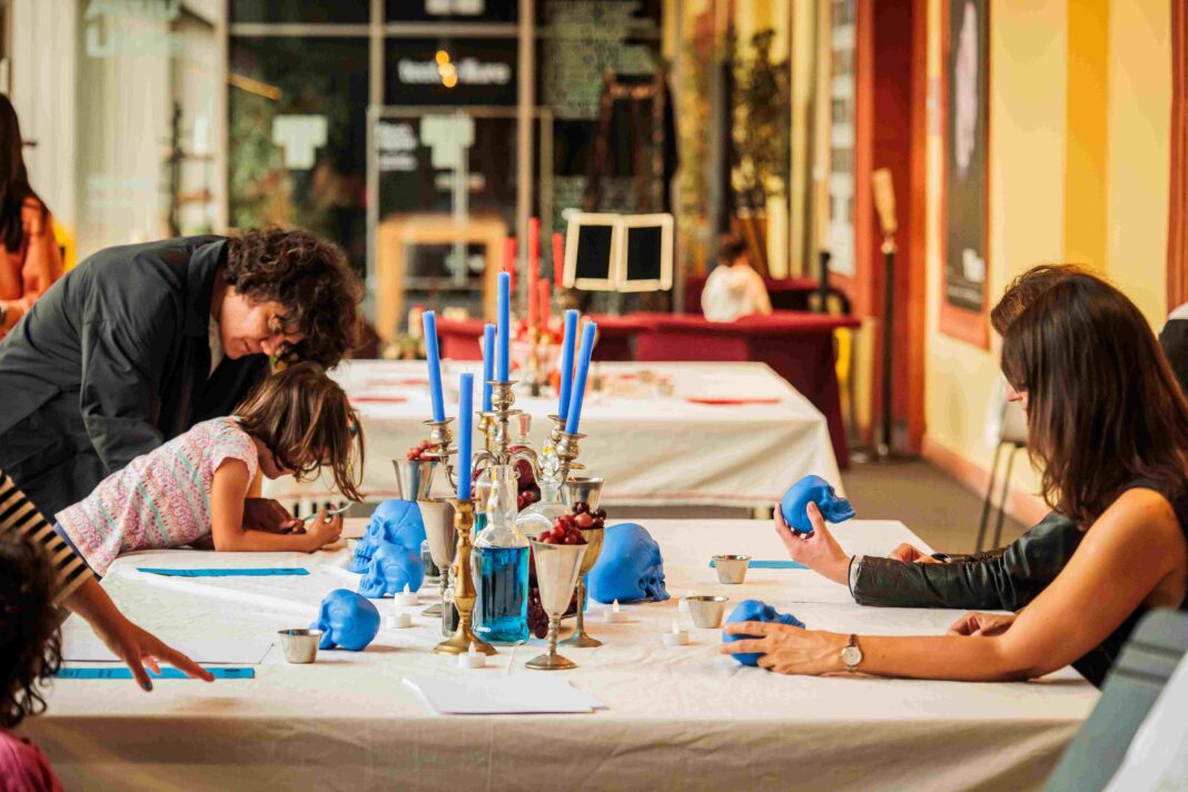Participants de diverses edats dibuixen al voltant d'una taula amb un canelobre amb espelmes blaves i cranis decoratius blaus durant un taller del festival Barcelona Dibuixa.
